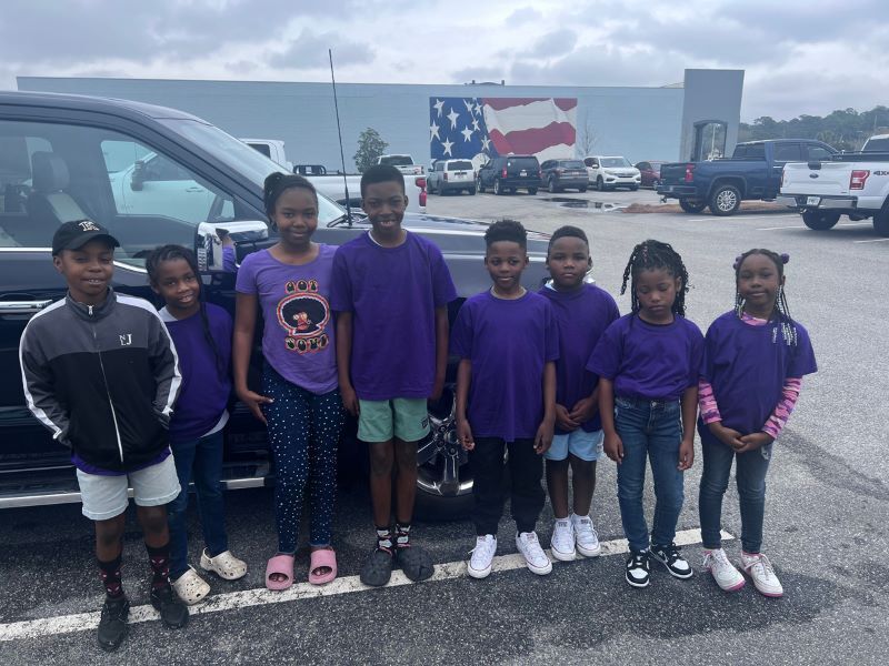 A group of children all wearing purple shirts while standing in front of a black car.